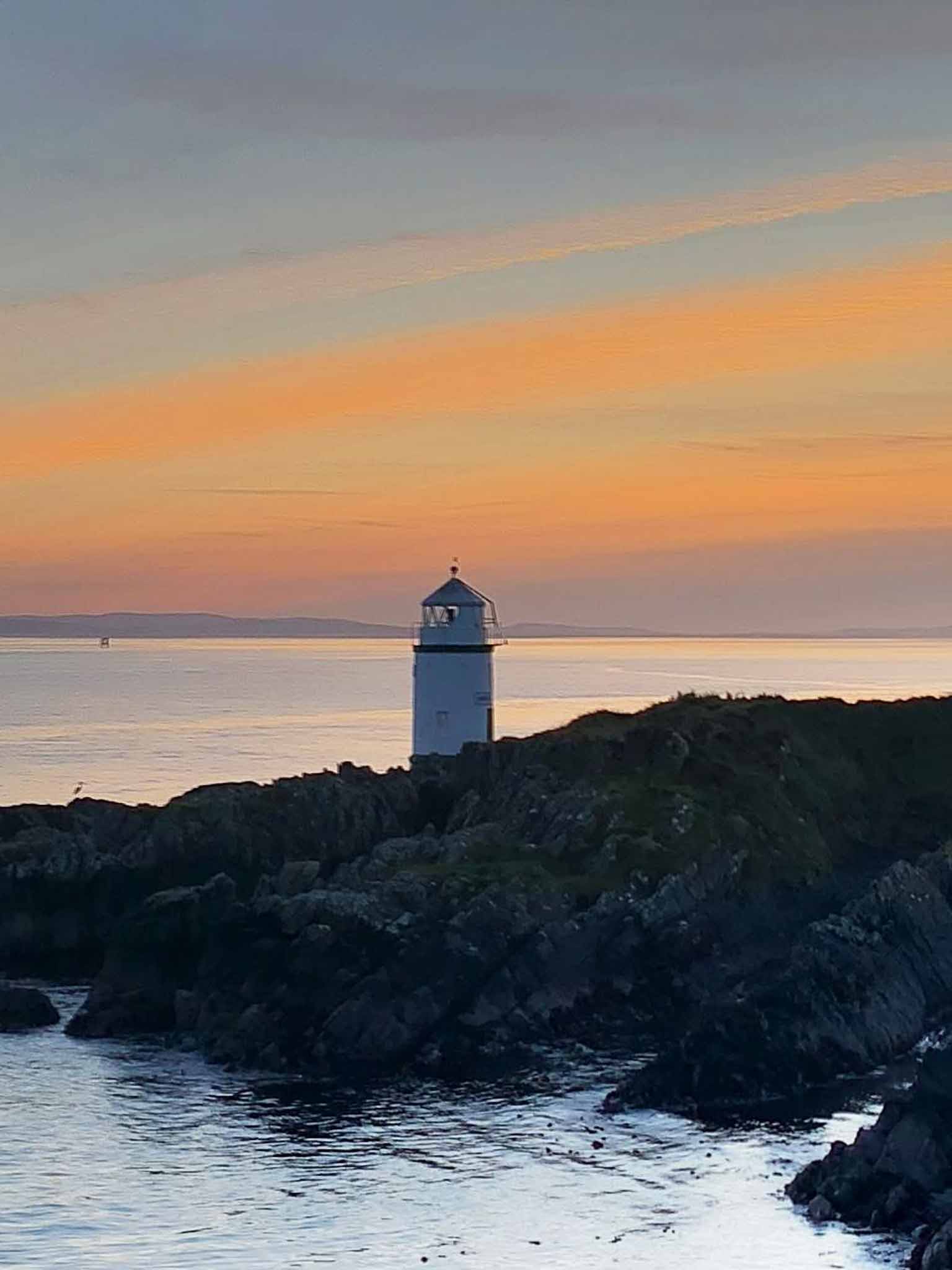 A white lighthouse above dark rocks beneath an orange evening sky